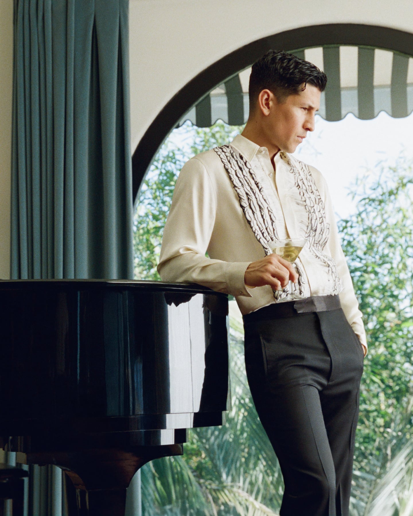 Actor Danny Ramirez is leaning on a piano at Amiri Chateau in a top and bottom made by the brand.
